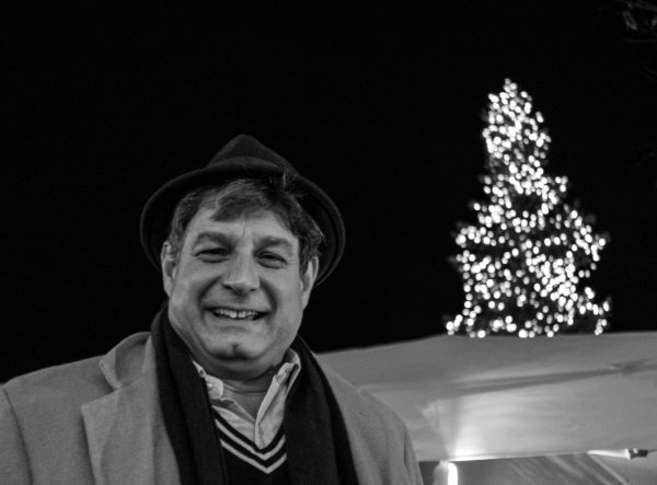 The coordinator of the tree lighting ceremony smiles joyfully. In the background to his right, the illuminated tree glistens. 